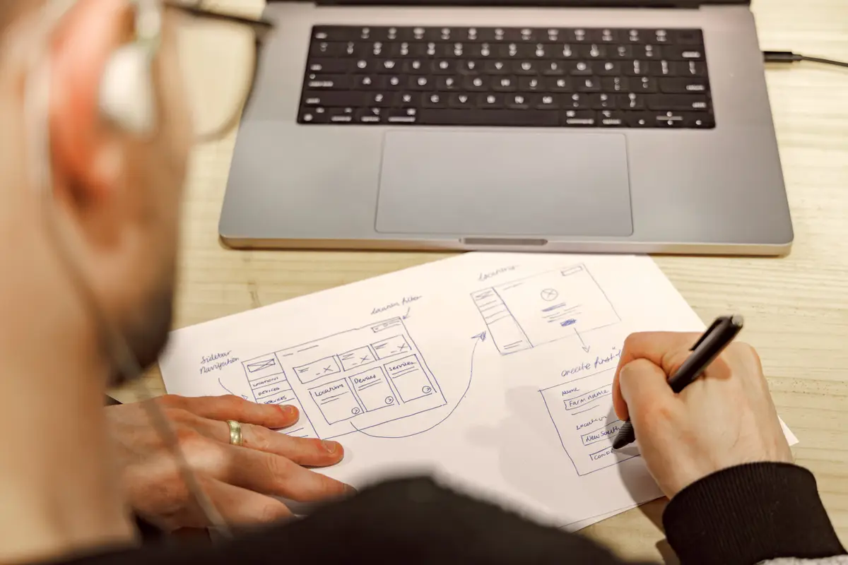 Person sketching app wireframes on paper beside a laptop