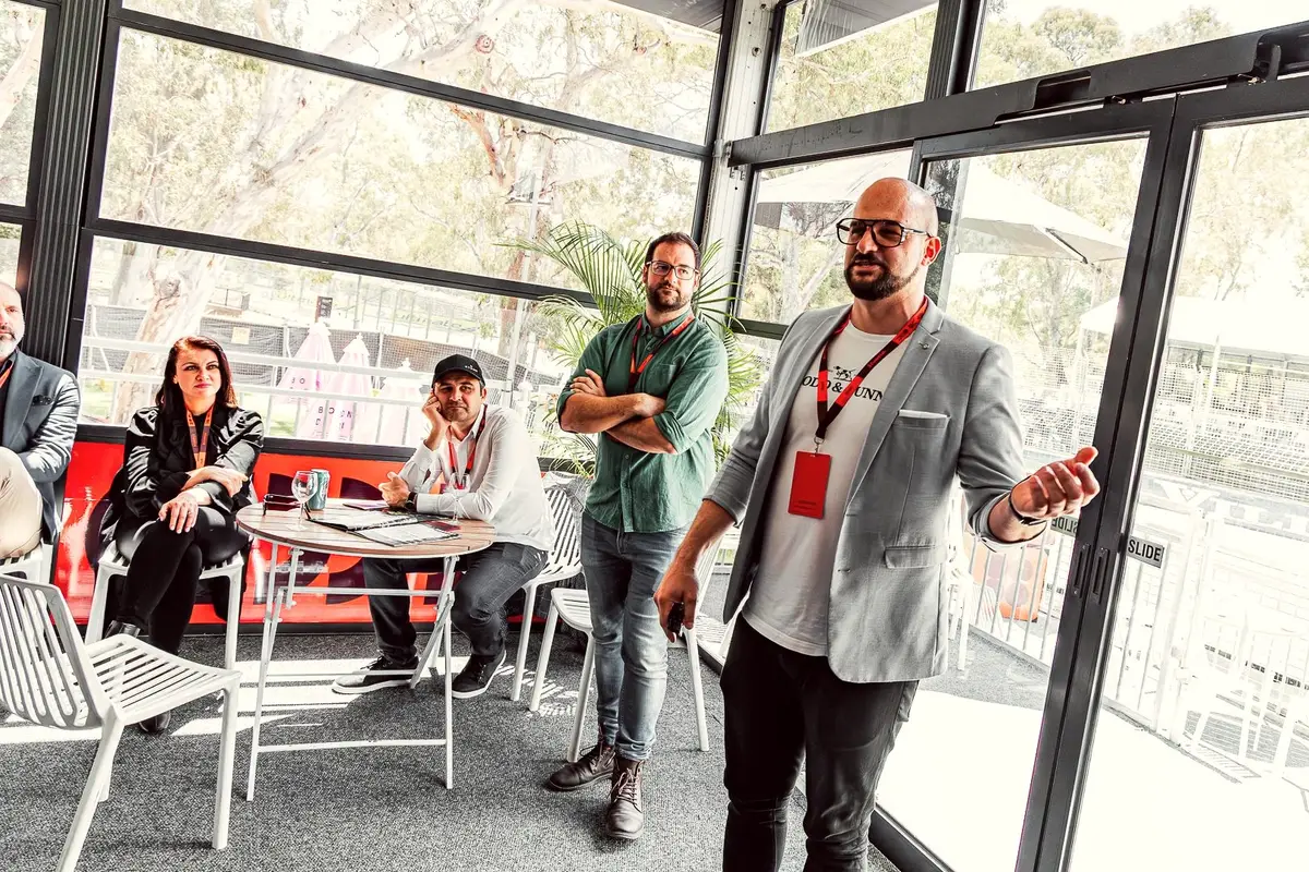 Man with a lanyard presenting to a small group in a sunlit glass-walled room