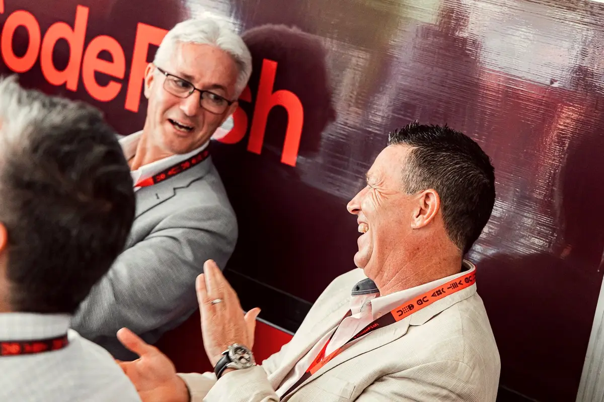 Three men laughing and talking in front of a CodeFish branded wall, wearing lanyards