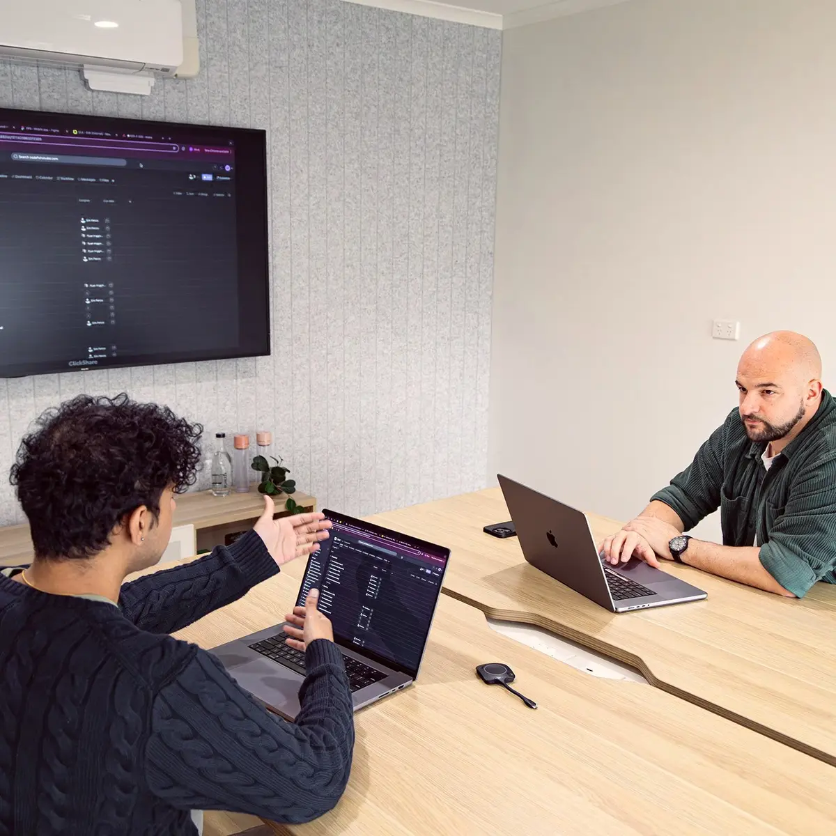 Two people discussing work on laptops in a meeting room with a wall screen