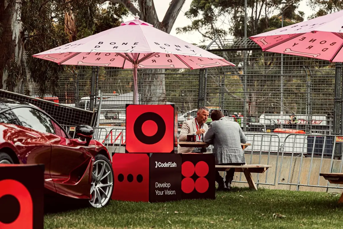 Two men talk at a picnic table beside CodeFish branded cubes and a red sports car