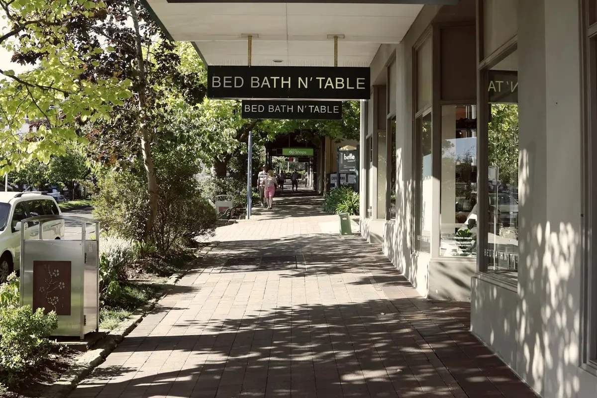 Tree-lined shopping street footpath under a Bed Bath N' Table store awning