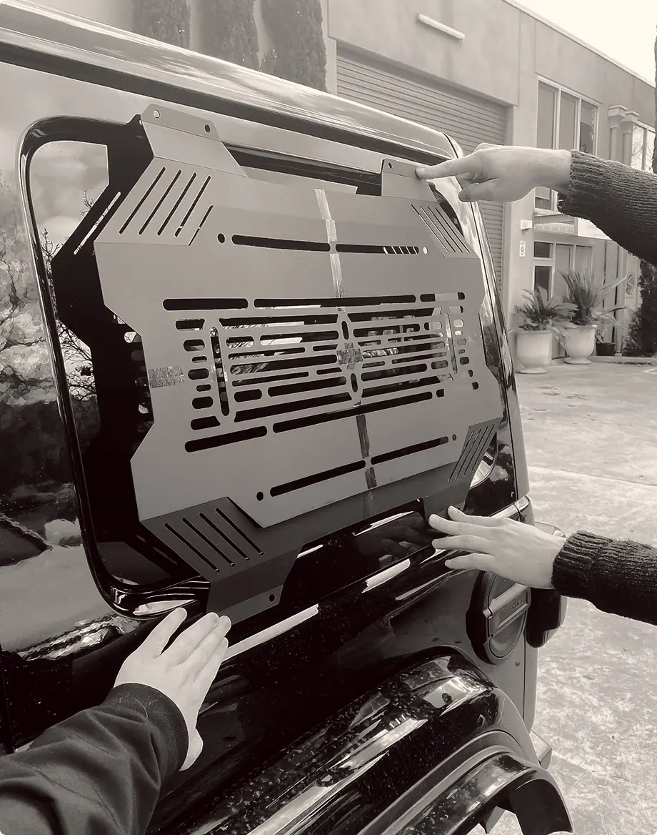 Two people holding a black storage panel in place over a vehicle rear window during a test fit