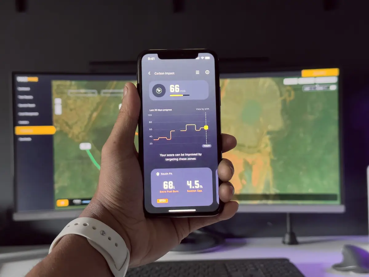 Hand holding a phone showing a carbon impact score app, with a mine site map on monitors behind