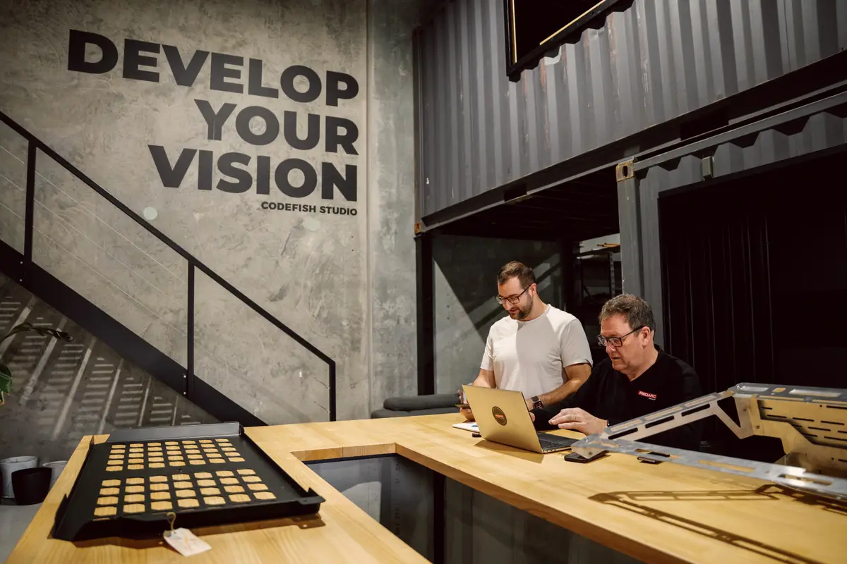 Two people working at a laptop beneath the CodeFish Studio 'Develop Your Vision' wall