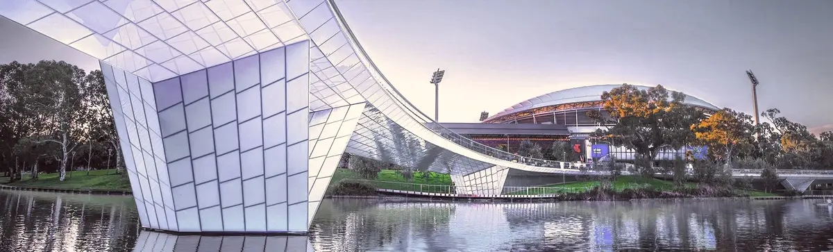 Glowing blue footbridge over the River Torrens with Adelaide Oval behind at dusk