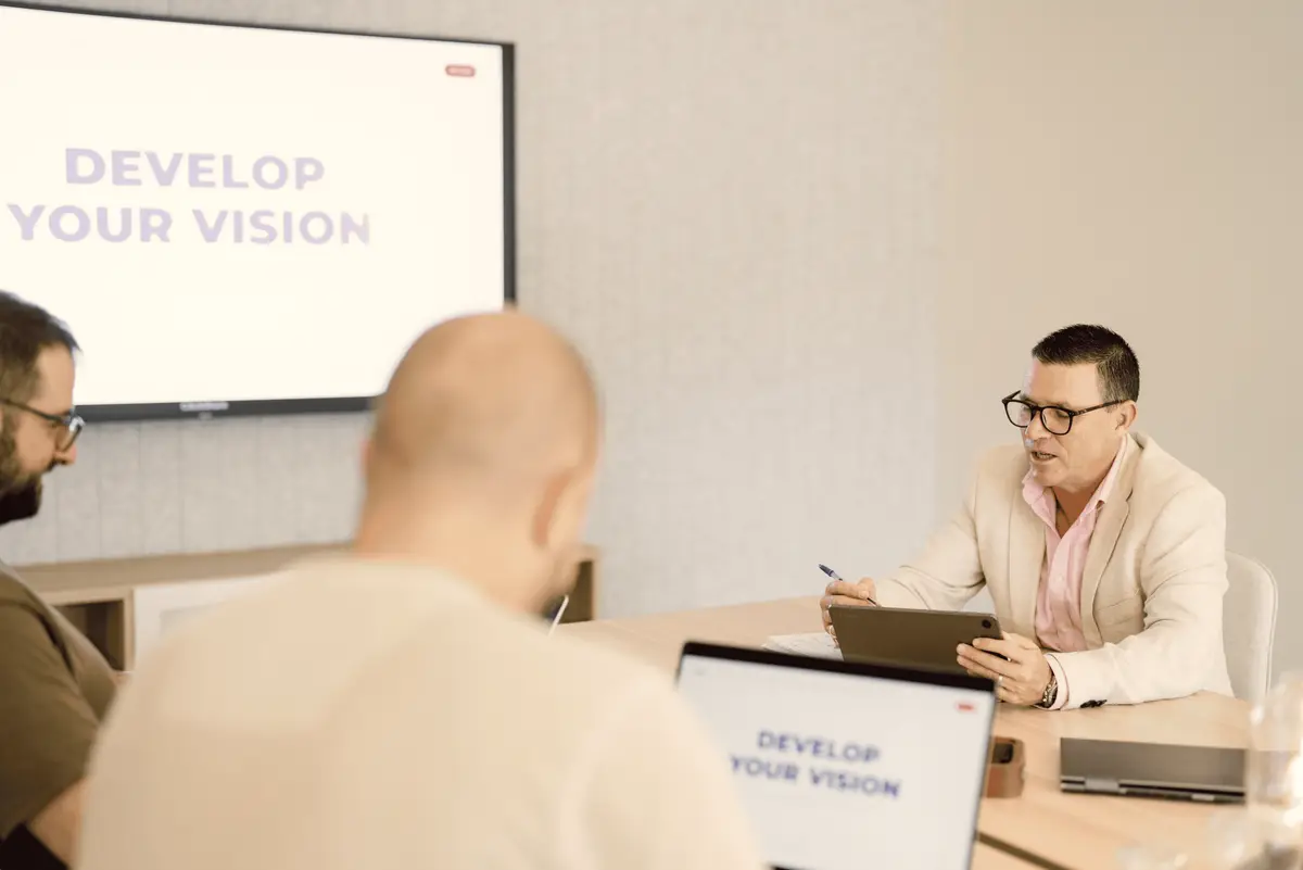 Meeting room discussion with a Develop Your Vision slide on the screen and a laptop